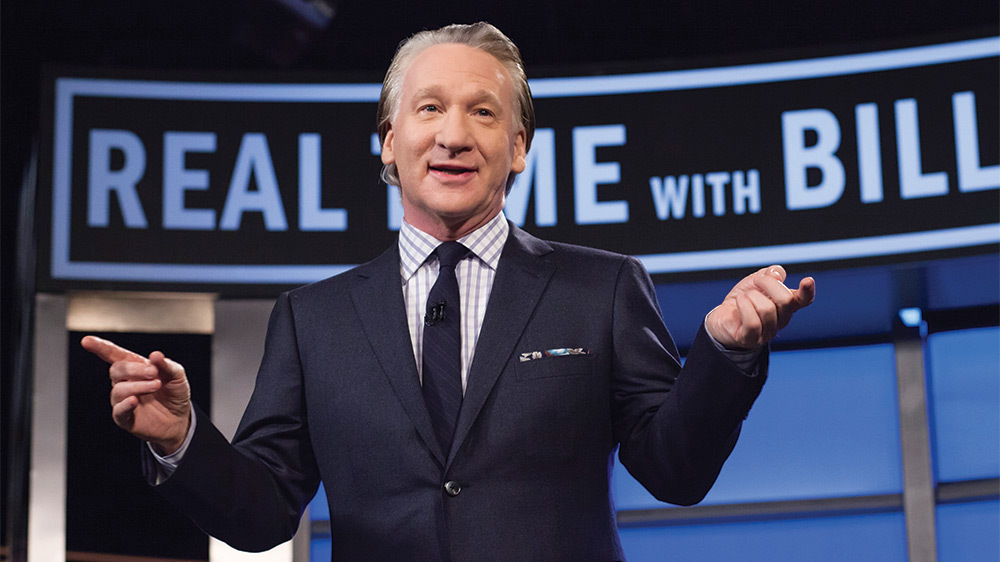 Comedian Bill Maher stands on his set with show graphics behind him.
